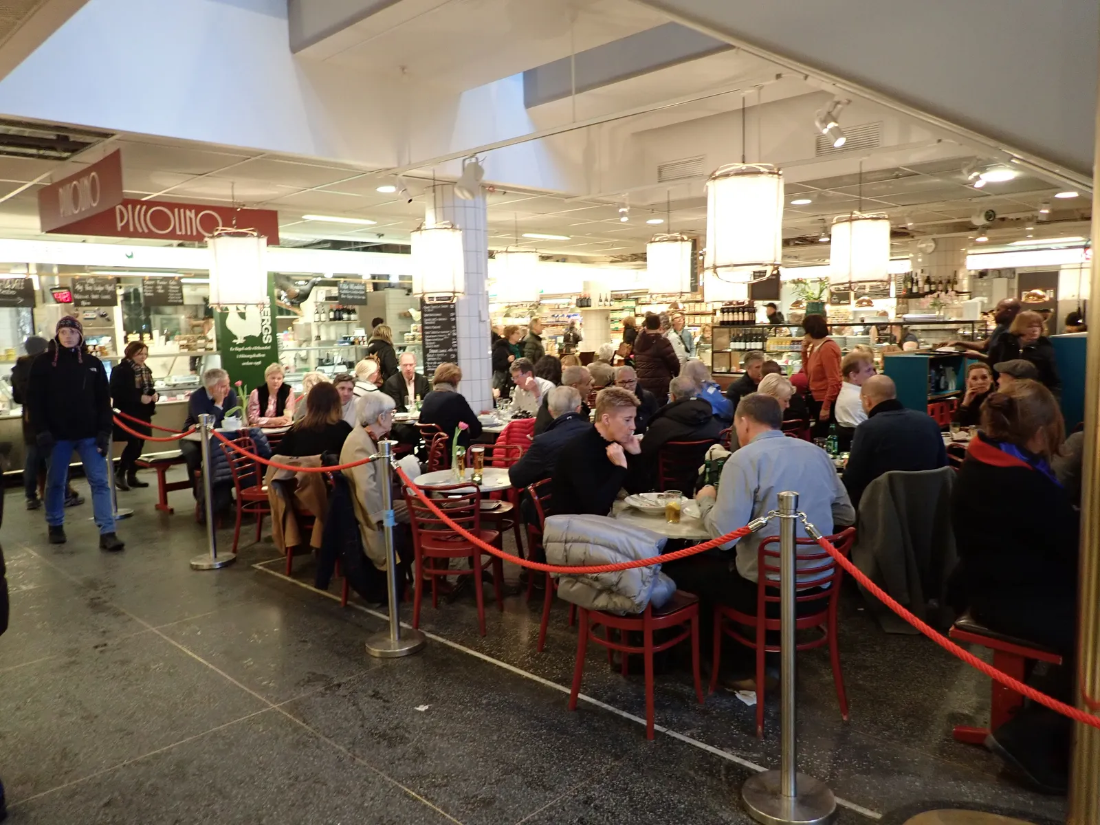 Inside Hötorgshallen food hall in Stockholm, people eating at red chairs and tables under hanging lamps with delicatessen and fish counters lining the aisle behind them