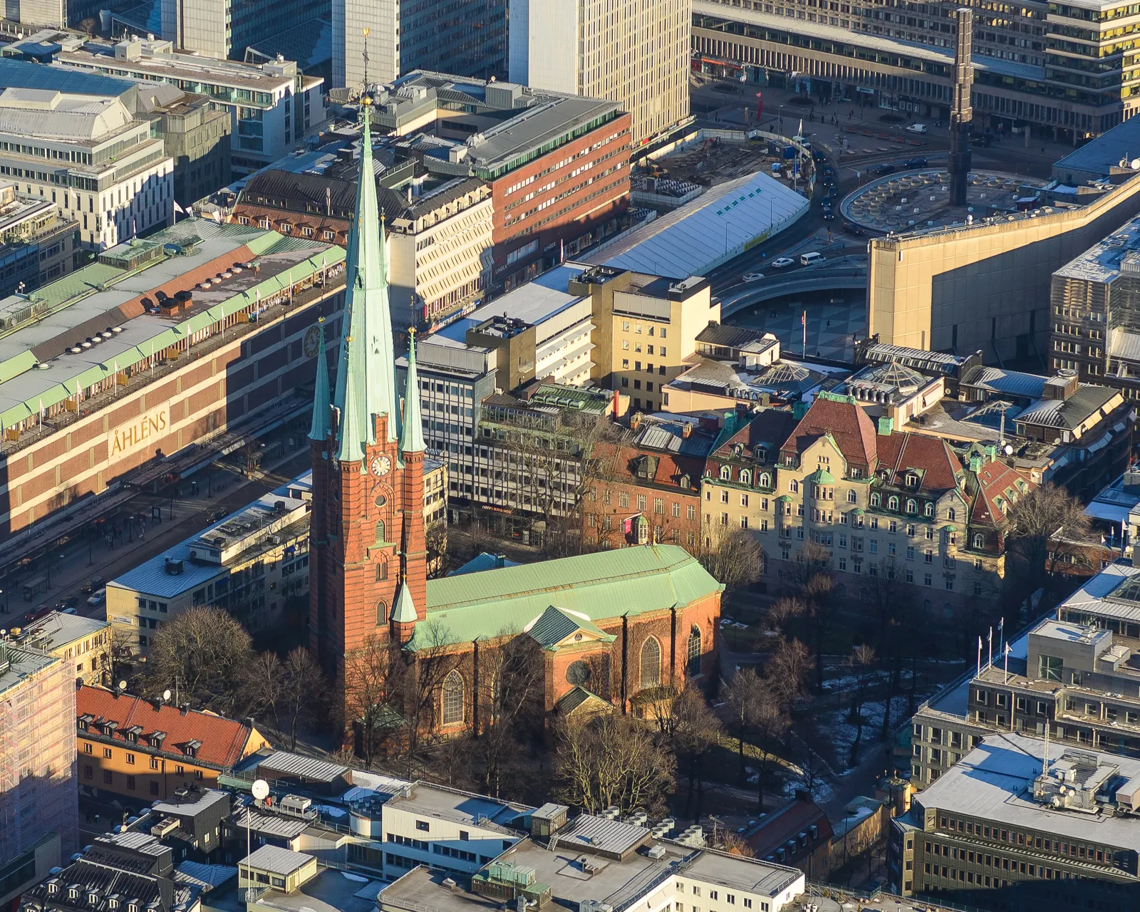 Klara Church in central Stockholm seen from above, its green copper spire and red brick nave standing in a walled churchyard of bare trees, hemmed in on every side by flat-roofed office blocks