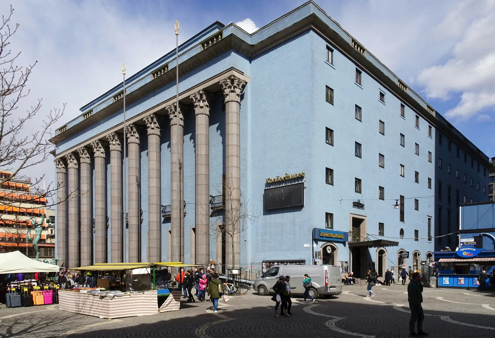 Stockholm Concert Hall on Hötorget, a pale blue block fronted by ten tall stone columns under a flat pediment, with green-canopied market stalls set out on the square in front of it