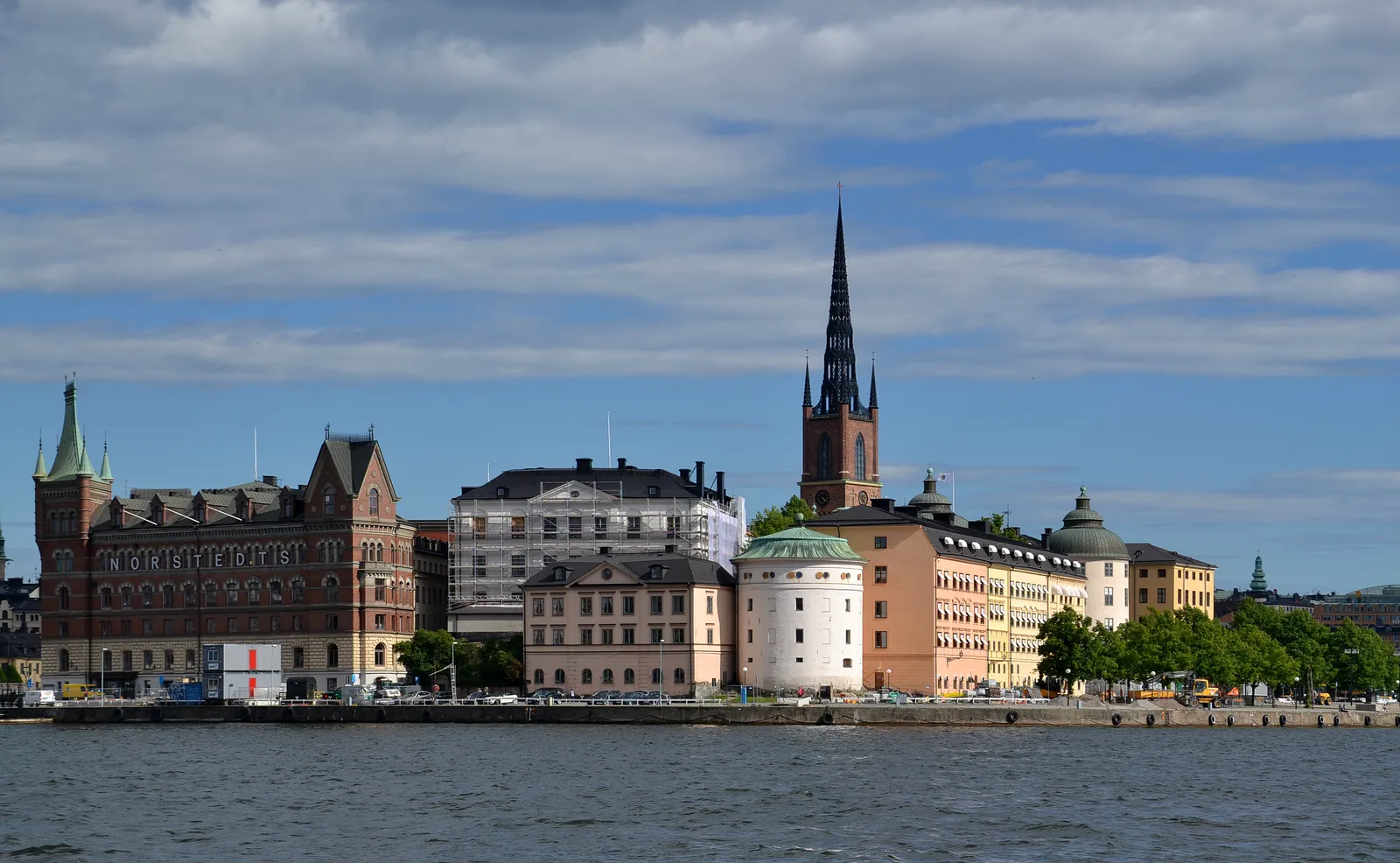 The island of Riddarholmen in Stockholm seen across the water, a row of pale 17th-century palace facades along the quay with the dark openwork cast-iron spire of Riddarholmen Church rising behind them