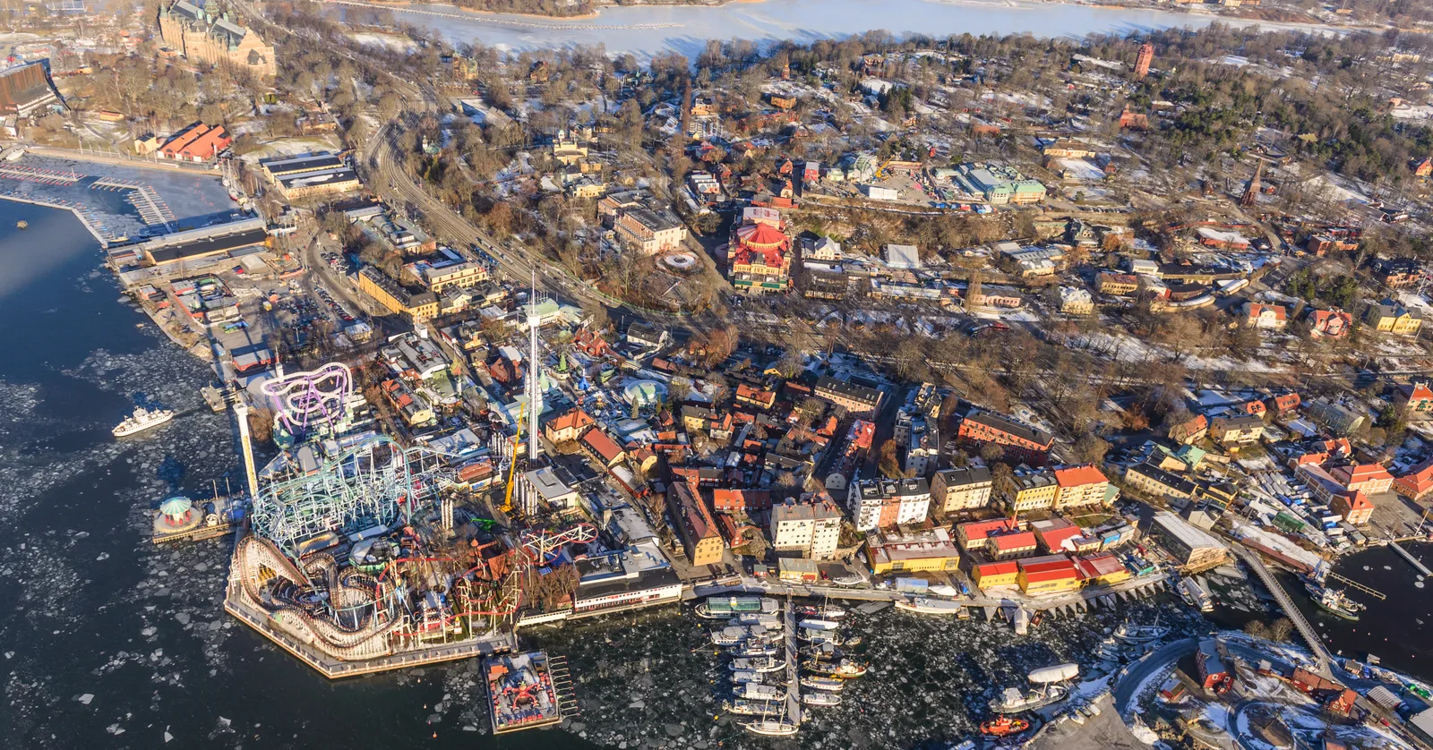 The western end of Djurgården in Stockholm from above in winter, an amusement park's rollercoaster track along the icy shoreline with wooden villas and bare trees covering the island behind it