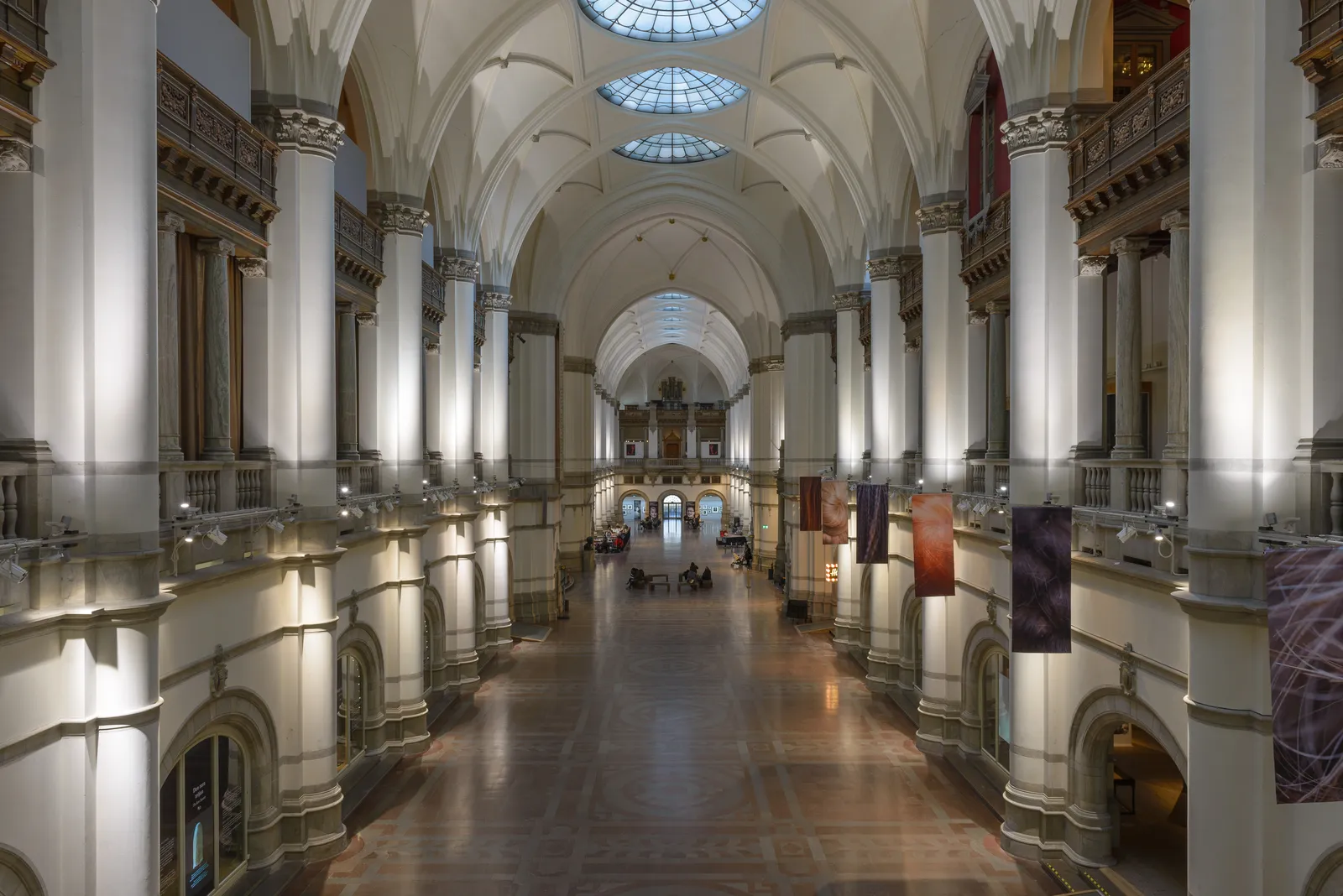The Great Hall of the Nordic Museum in Stockholm, an enormous vaulted room with white columns and balconied galleries running down both sides towards a lit figure at the far end, glass domes in the roof above