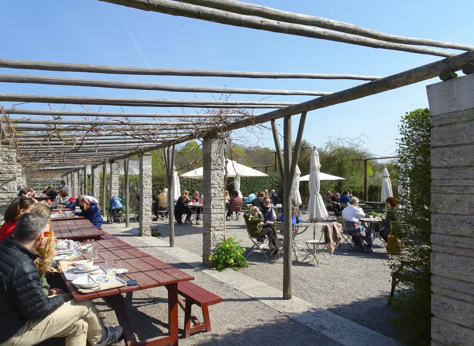 The terrace at Rosendal's garden in Stockholm, people eating at long wooden trestle tables under a bare timber pergola with stone paving, parasols and planted beds around them