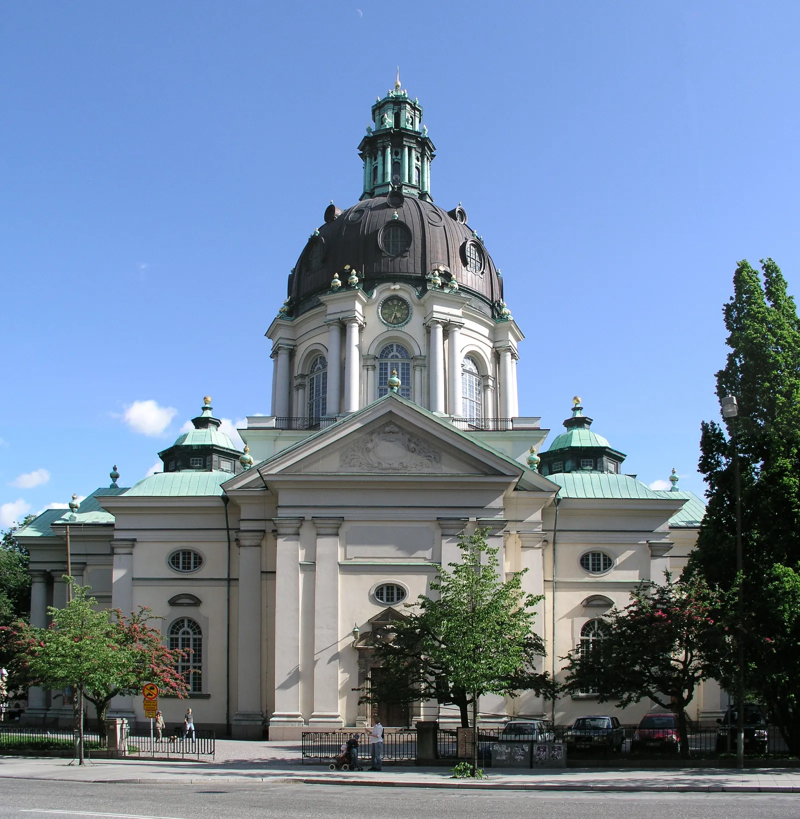 Gustaf Vasa Church in Stockholm under a blue sky, a pale stone building with a columned portico and a dark dome topped by a green copper lantern, trees along the street in front of it
