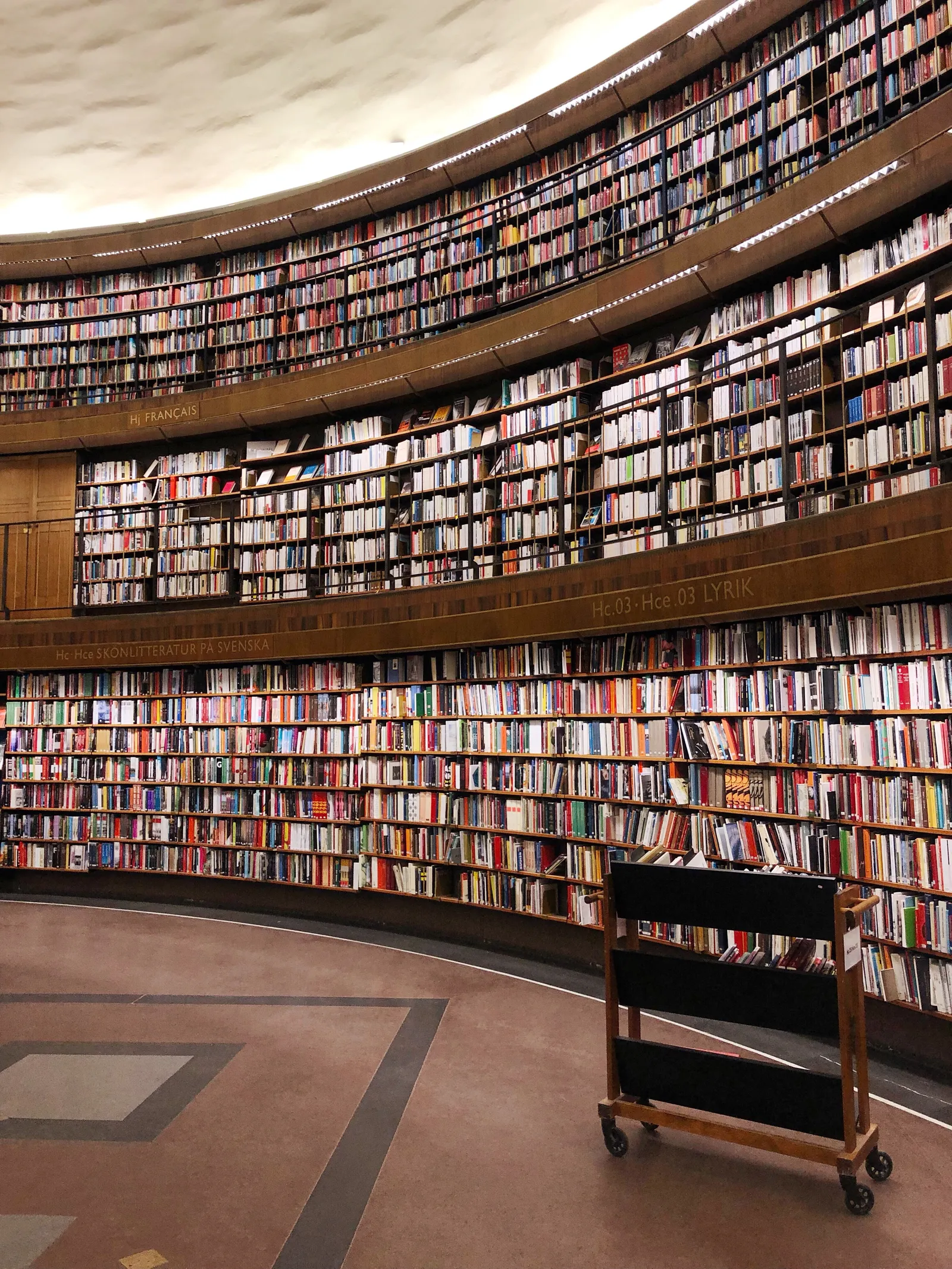Inside the round reading room of Stockholm Public Library, three curved tiers of open bookshelves packed with spines running all the way around the wall under a white ceiling, with a wooden book trolley on the floor