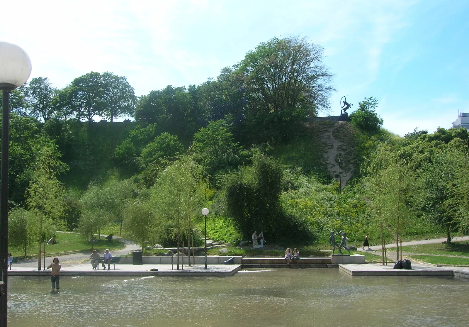The wooded hill of Observatorielunden in Stockholm rising behind a shallow public water basin, with people sitting on the steps around the water and a dark statue silhouetted on the rock face at the top