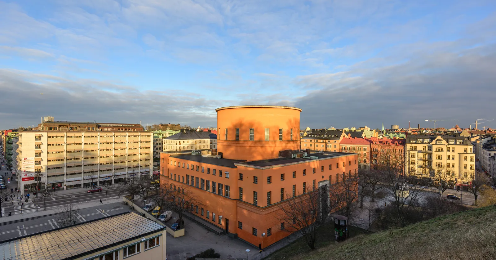 Stockholm Public Library seen from above in low winter sun, a tall orange cylinder rising out of a square orange block, with the apartment rooftops of the surrounding district spread out behind it