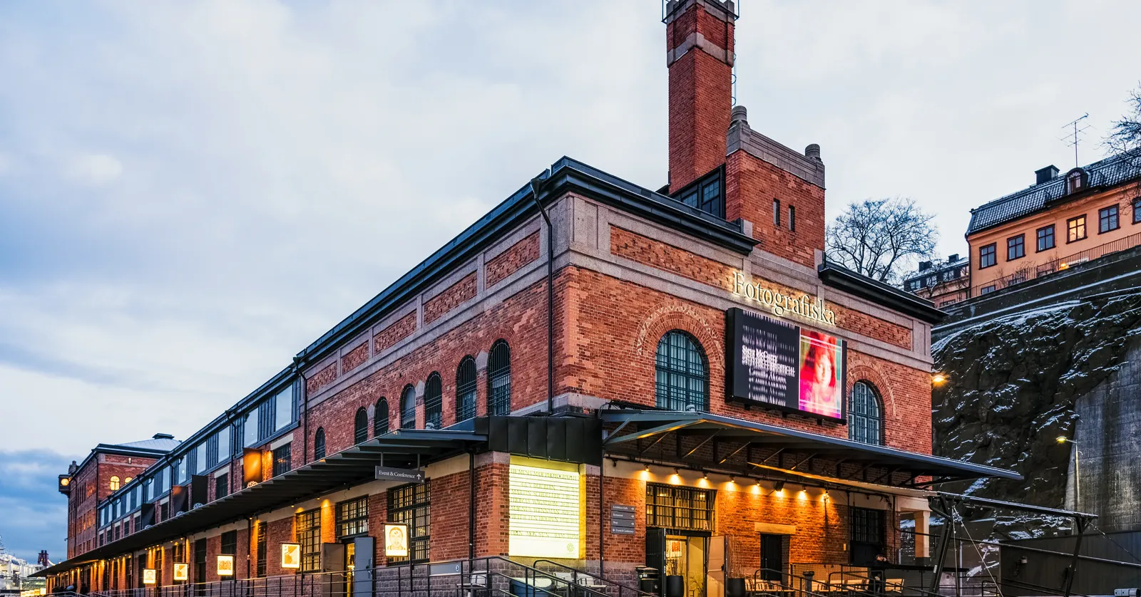 The brick customs house on the Stockholm quayside that houses the Fotografiska photography museum, a long arcaded red-brick building with a square chimney tower, the museum's name lettered along the side and its entrance lit at dusk