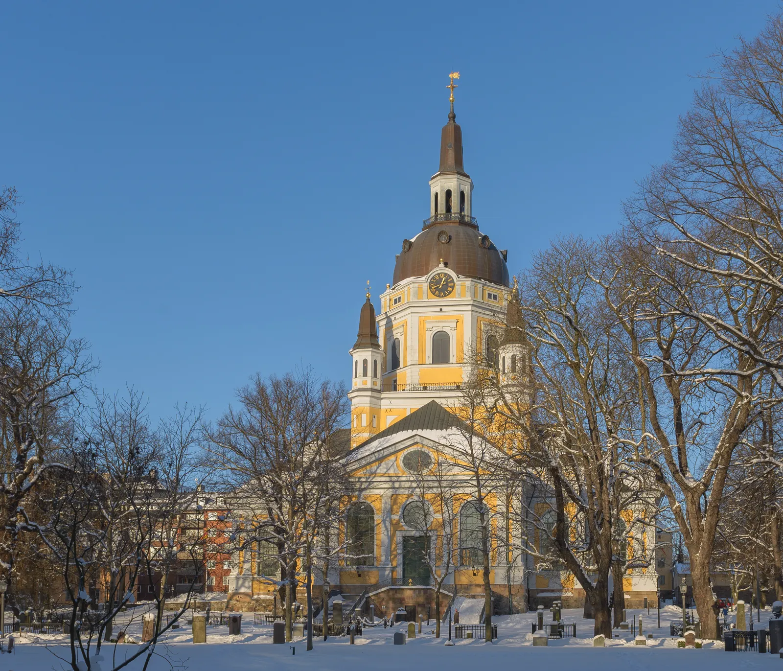 Katarina Church on Södermalm in Stockholm under snow, a pale yellow building with a dark octagonal tower and dome topped by a gilded finial, standing among bare trees in its graveyard