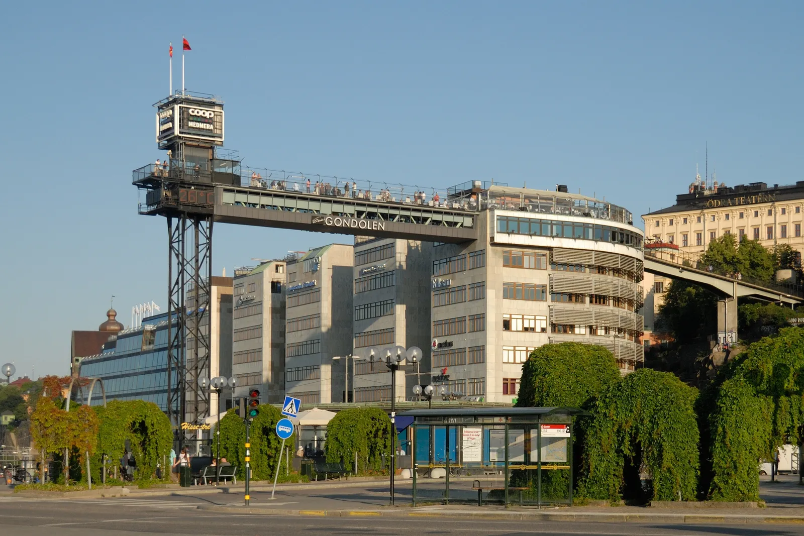 The Katarina lift in Stockholm, a slim steel tower carrying a high walkway across to the roof terrace of an office building, with people standing along the walkway railing above the street