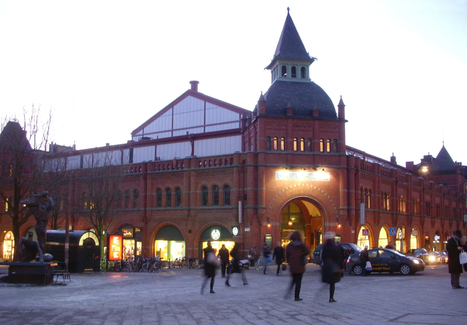 Östermalms Saluhall in Stockholm at dusk, a red brick market hall with arched windows, a glazed upper gable and a pointed corner turret, with people crossing the square in front of its lit entrance