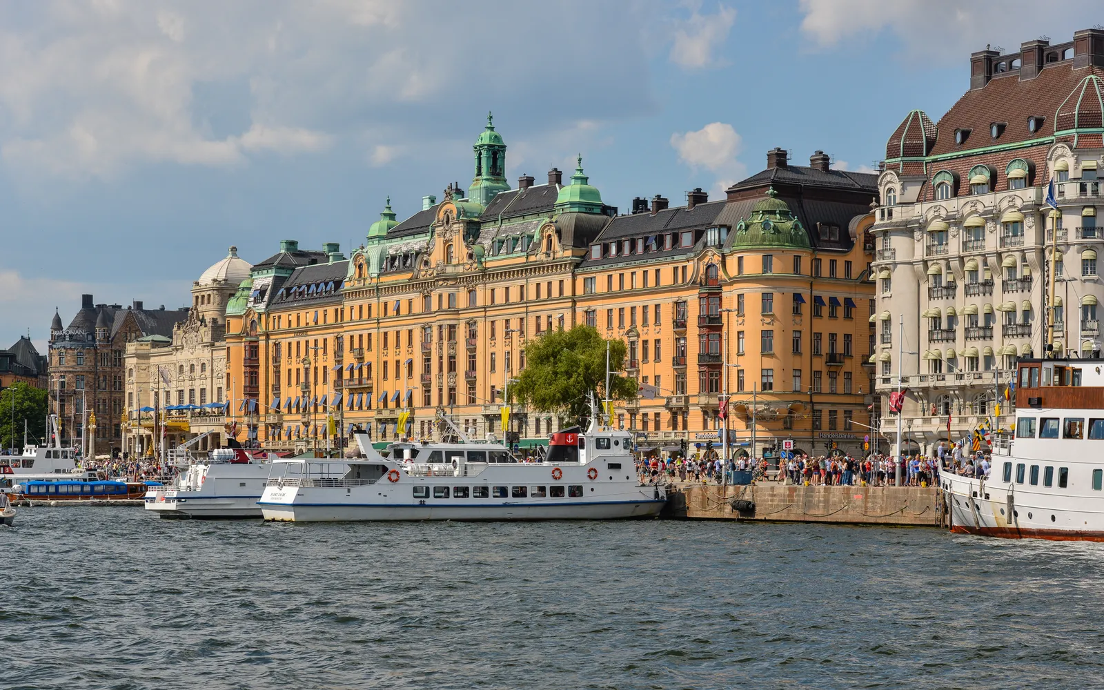 Strandvägen in Stockholm seen from the water, a row of ornate turn-of-the-century apartment facades with green copper roofs above a quay lined with white passenger boats