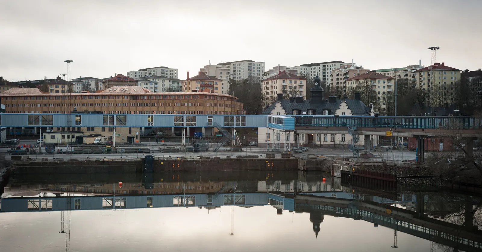 The Värtahamnen ferry terminal in Stockholm seen across the harbour basin, low blue and timber-clad terminal buildings along the quay with the apartment blocks of the Gärdet district rising on the slope behind them