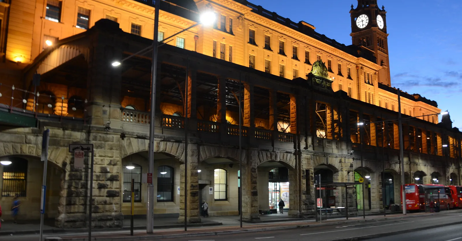 The sandstone frontage of Central Station in Sydney at dusk, its arcaded lower storey and long upper floors lit up, with the clock tower rising at the far end
