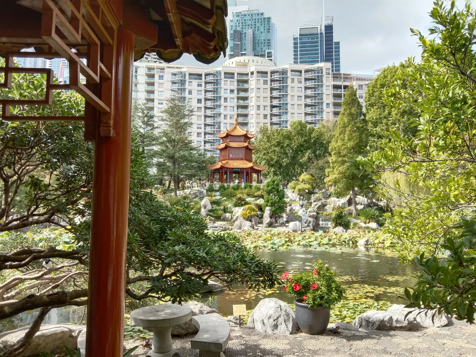 A red pagoda on a rise above the lake in the Chinese Garden of Friendship in Sydney, framed by a pavilion post and trees, with apartment towers behind