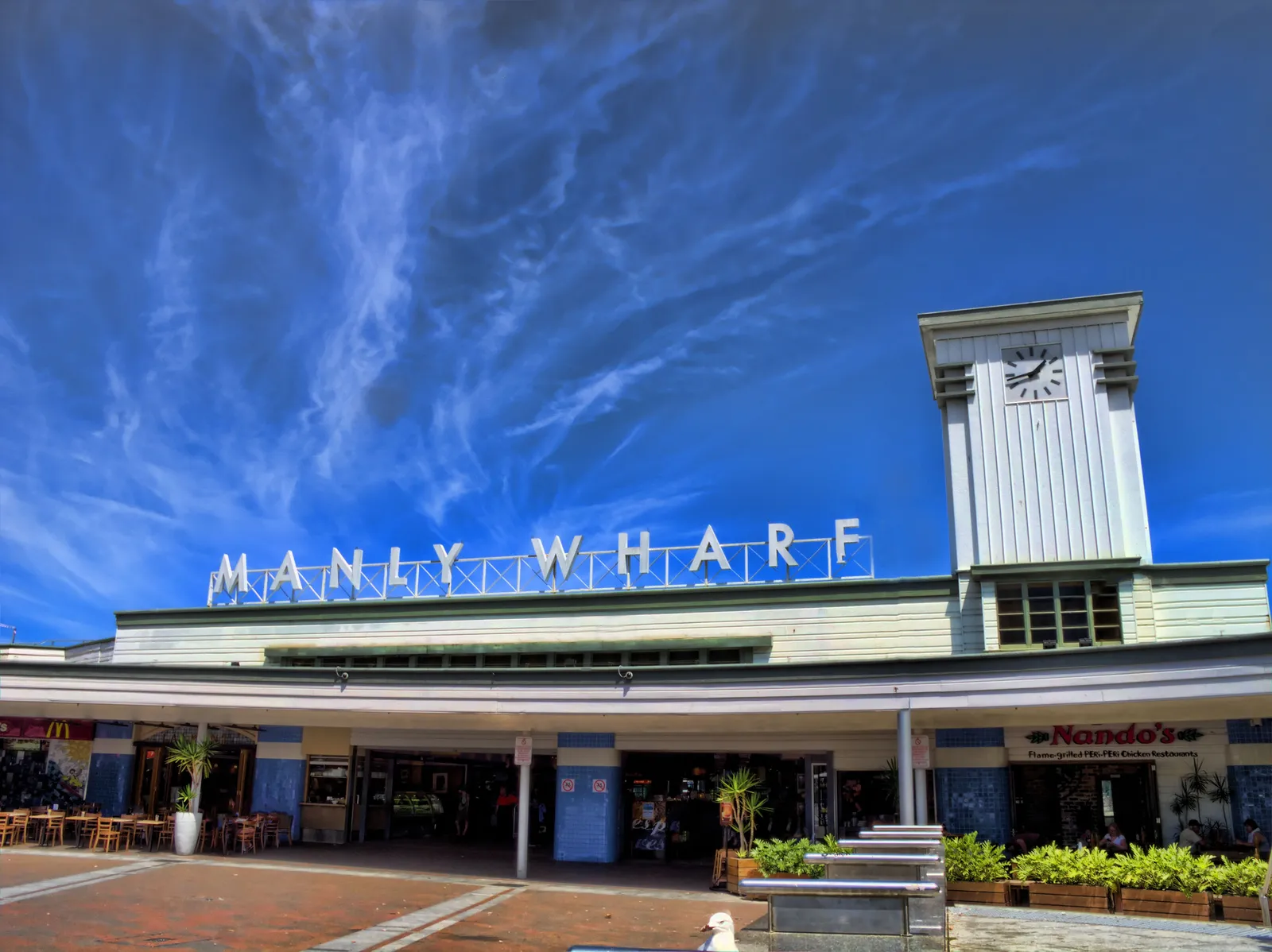 The white Manly Wharf building on Manly Cove, its clock tower and lettered facade above a paved forecourt and cafe tables