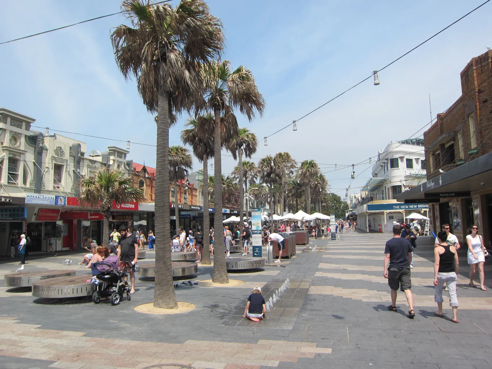 The Corso in Manly, a paved pedestrian street lined with palm trees, shopfronts and benches, with people walking toward the ocean end