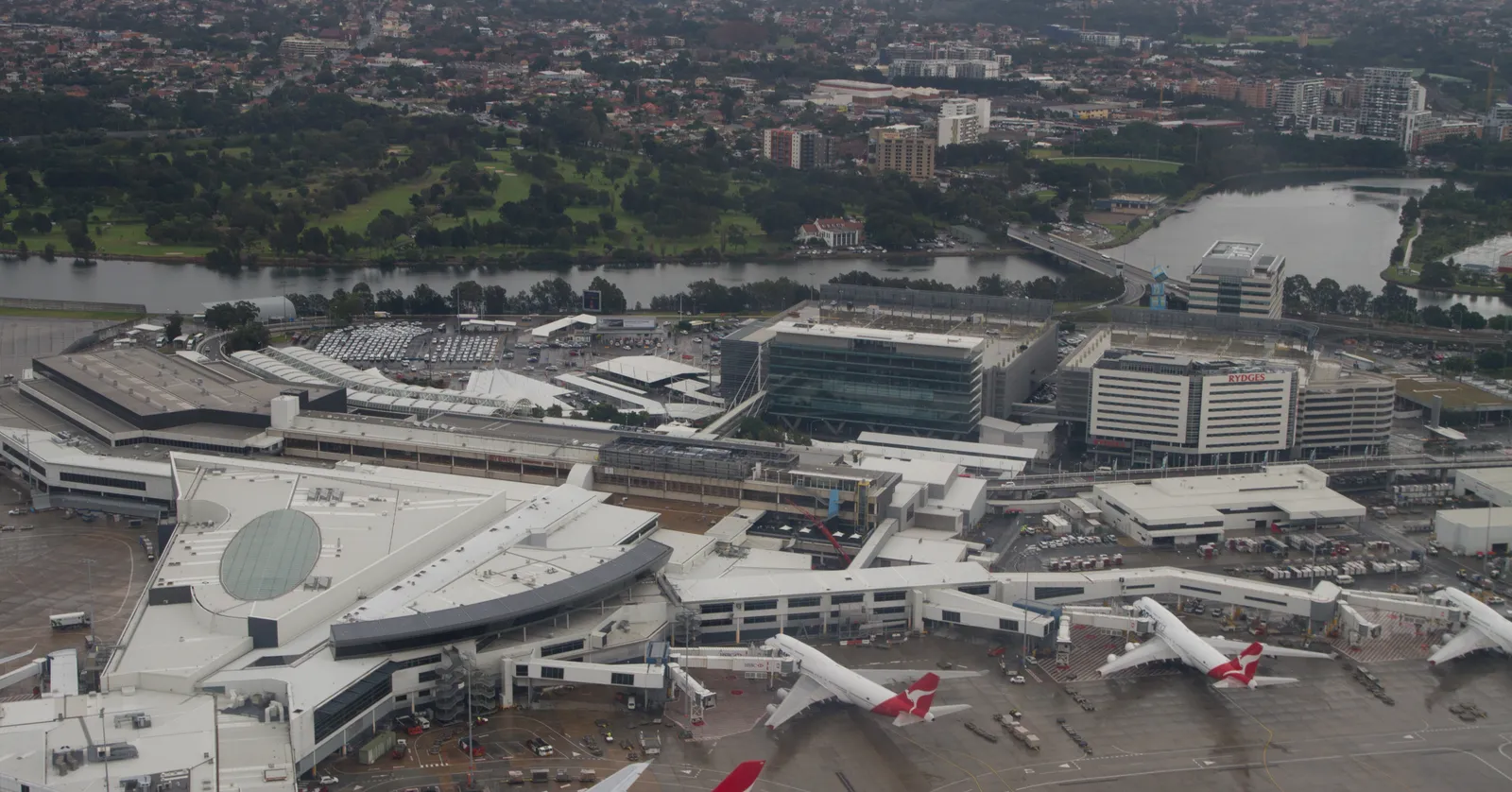 Sydney Airport's Terminal 1 seen from the air, with Qantas jets at the gates, the terminal car parks and hotel behind, and the Cooks River curving past beyond