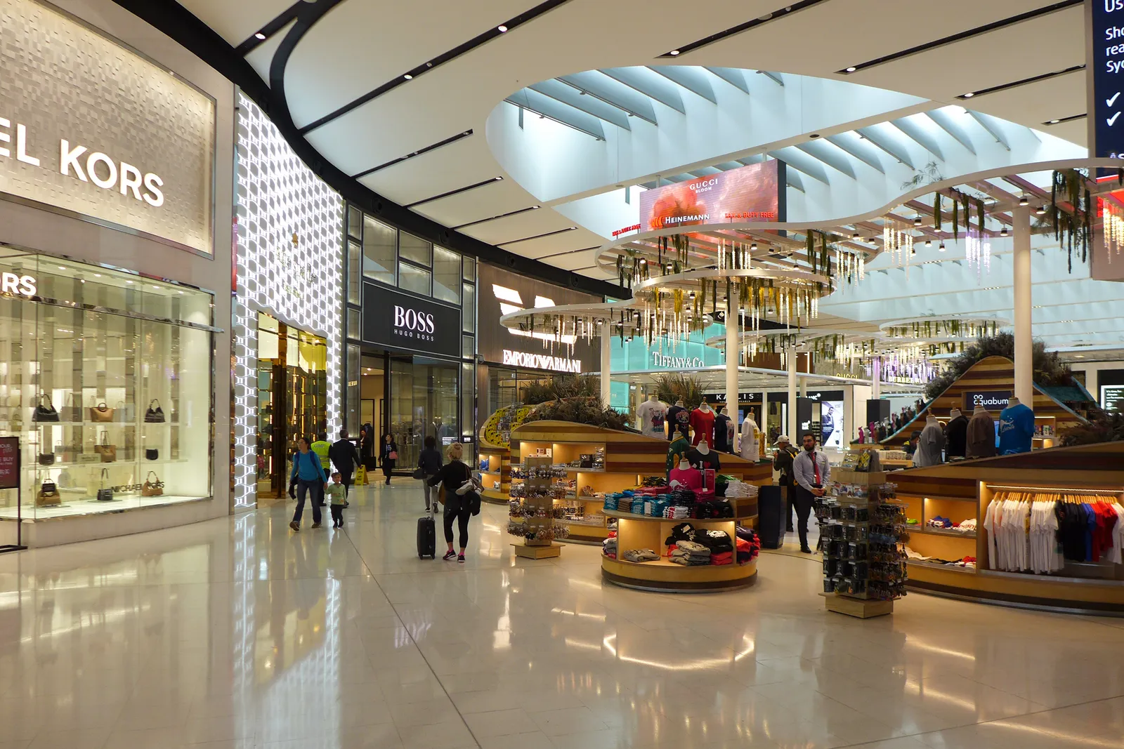 The airside shopping hall in Sydney Airport's Terminal 1 past security, with fashion shopfronts on the left and island counters under a curved white ceiling