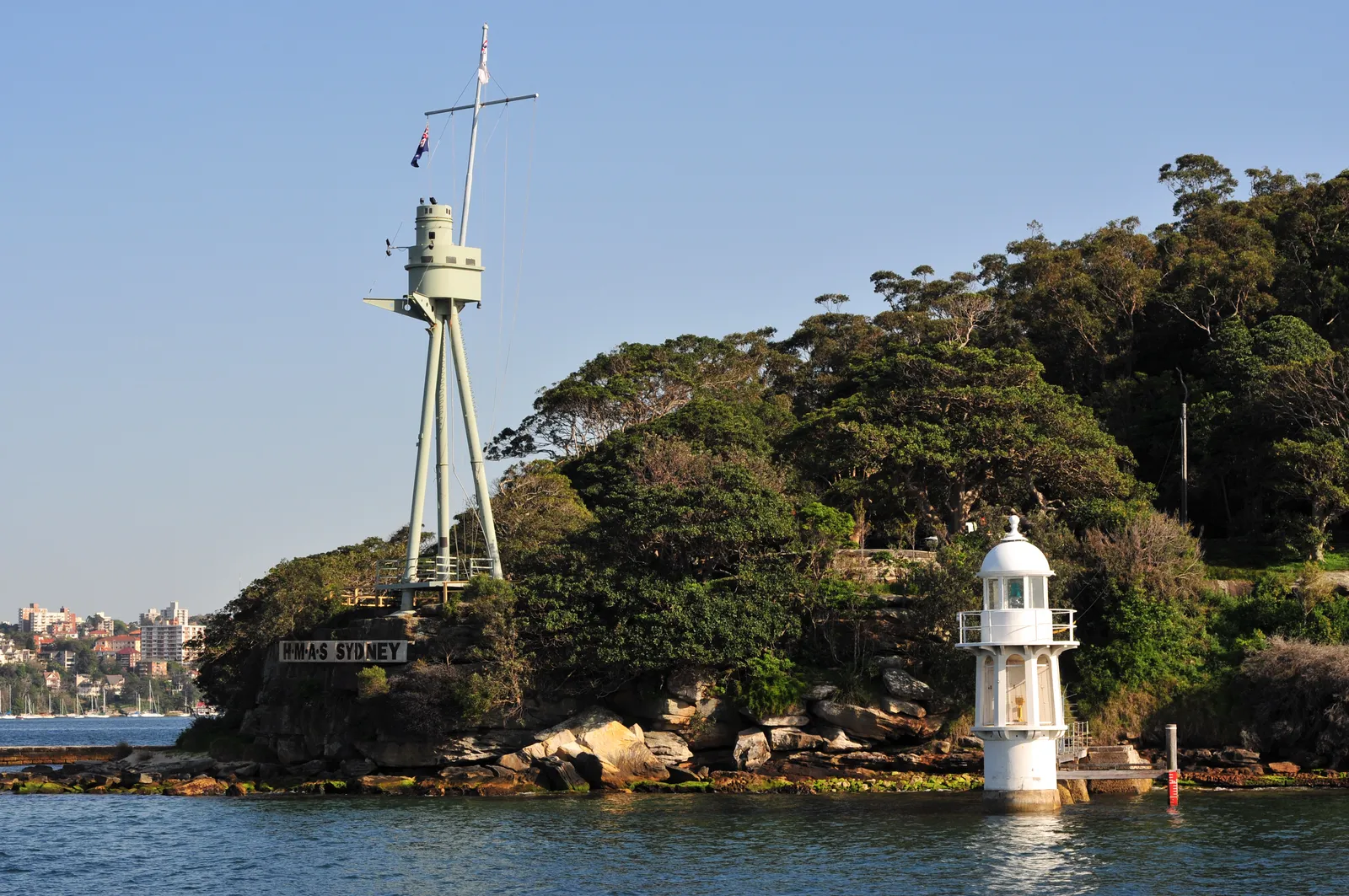 Bradleys Head in Sydney Harbour, the small white lighthouse on the rocks below the tall grey foremast of HMAS Sydney on the wooded headland