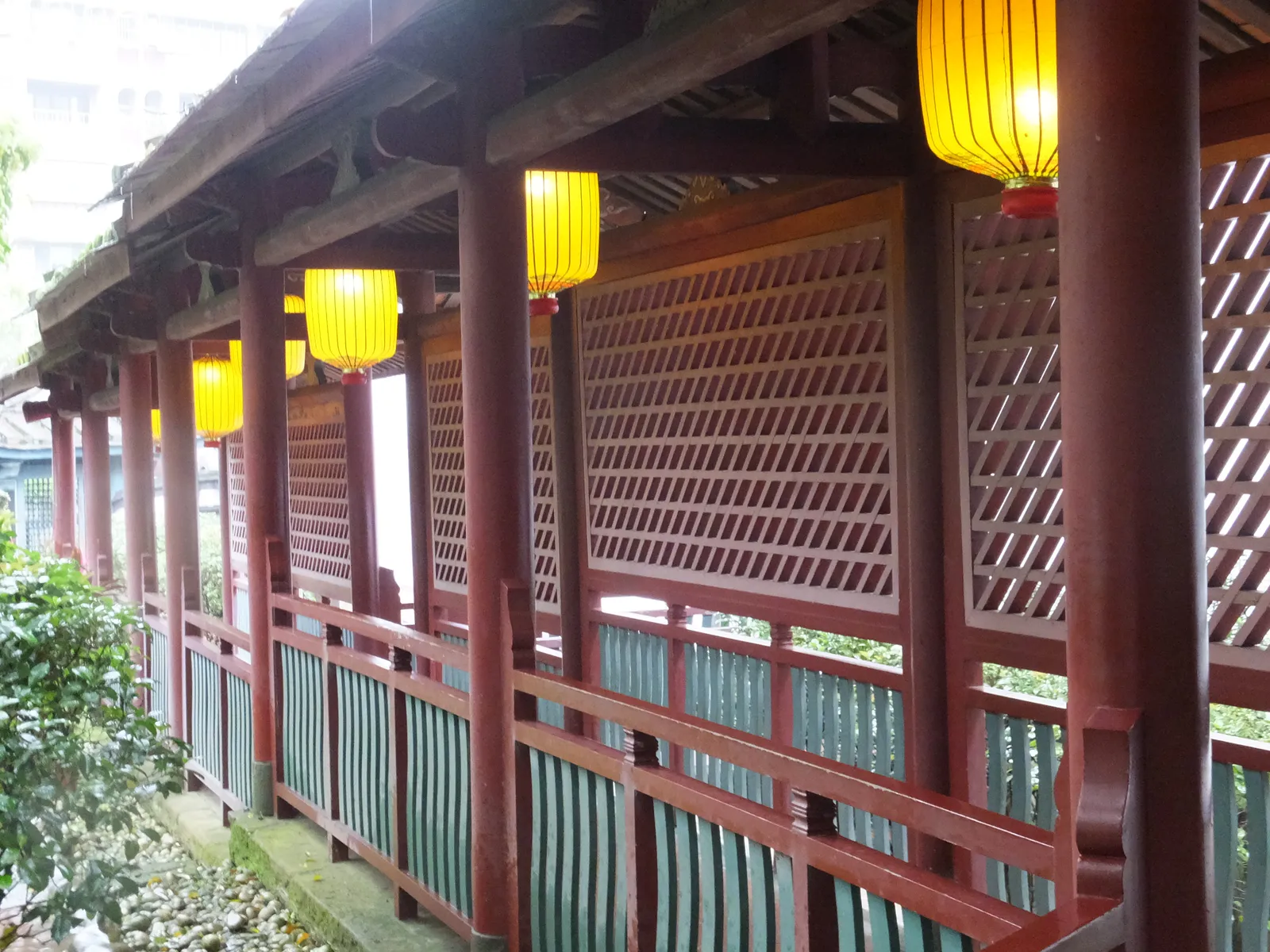 A red-painted covered walkway in the Lin Family Mansion and Garden in Banqiao, hung with yellow lanterns and screened by latticed wooden panels, with greenery beyond
