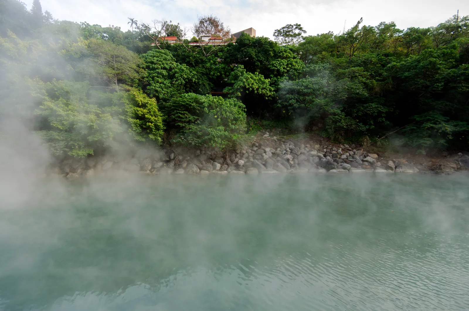 Steam rising off the milky green water of Thermal Valley in Beitou, Taipei, with a rocky bank and dense green hillside behind it