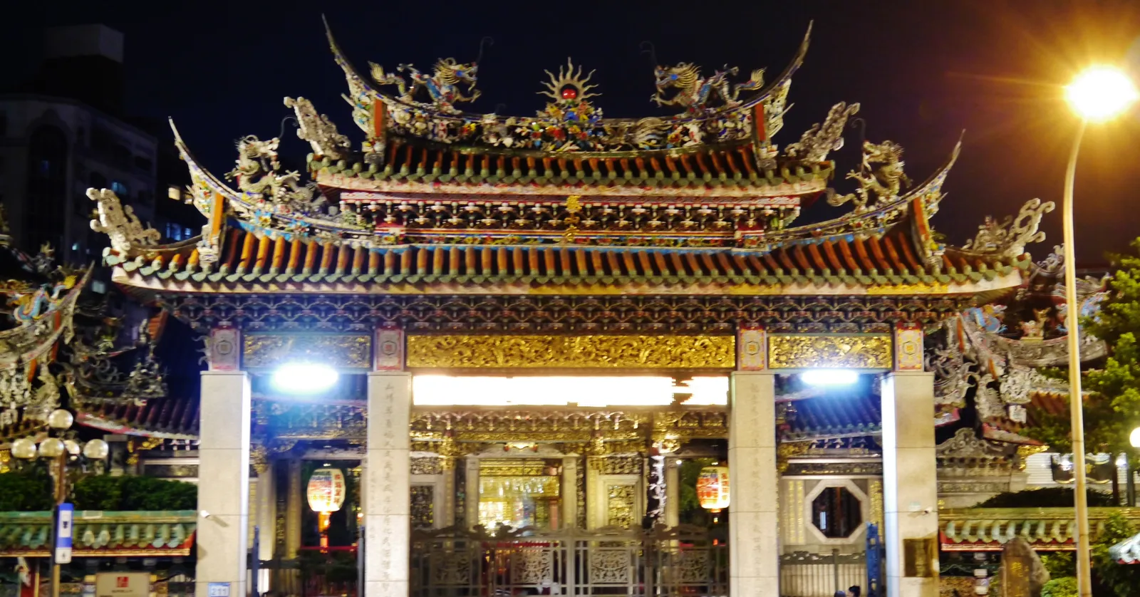 The front gate of Longshan Temple in Taipei lit up at night, its swallowtail roof crowded with ceramic dragons and figures above rows of red lanterns