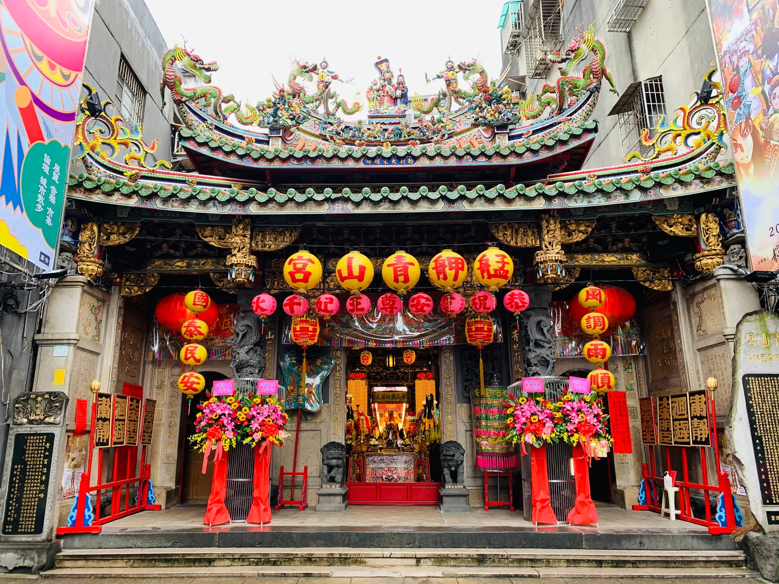 The front of Bangka Qingshan Temple in Taipei, hung with rows of red lanterns under an ornate tiled roof, with flower stands set out on either side of the doors