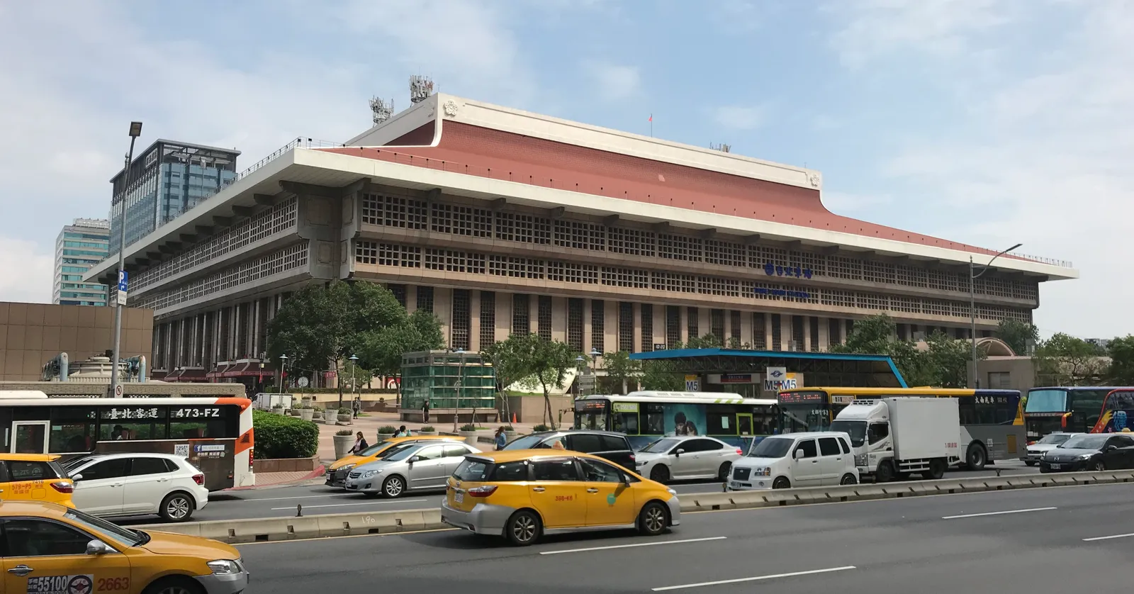 Taipei Main Station seen from across Zhongxiao West Road, a wide 1980s building with a sweeping red-tiled roof, buses and taxis passing in front of it