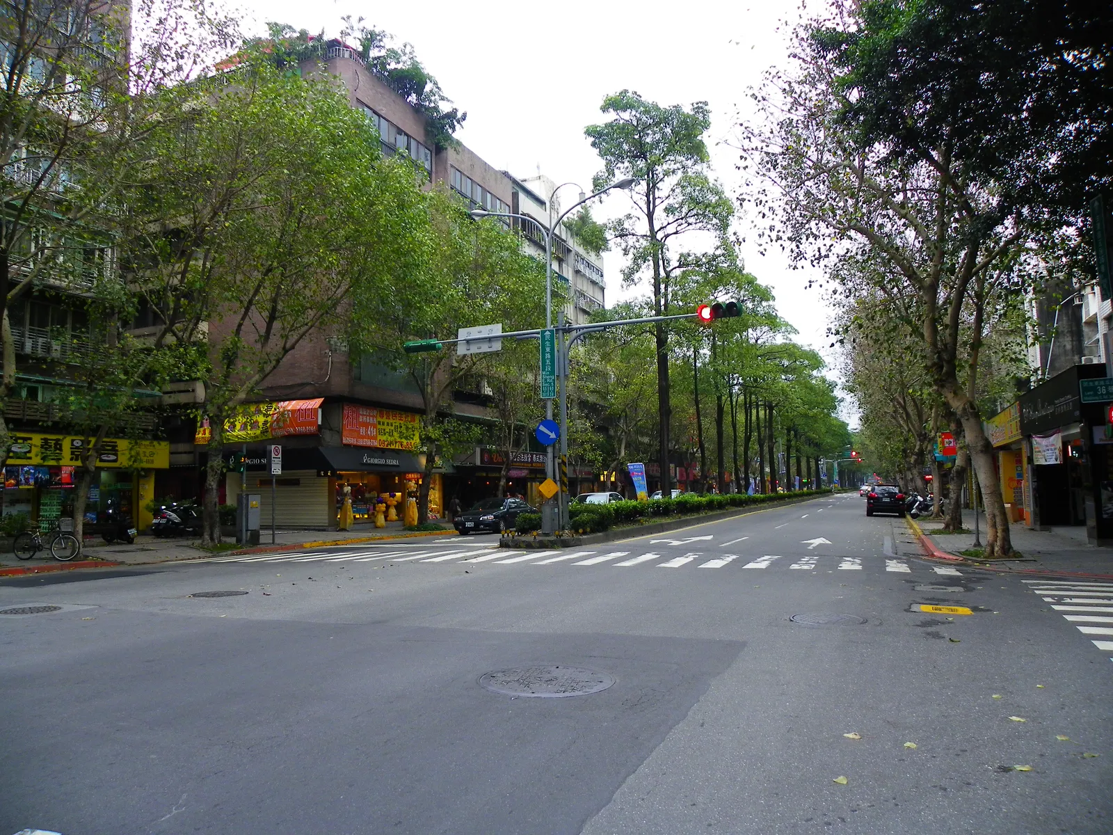 A wide tree-lined avenue in Taipei's Minsheng Community, low shopfronts along the left side and a long row of mature trees running away down the middle of the road