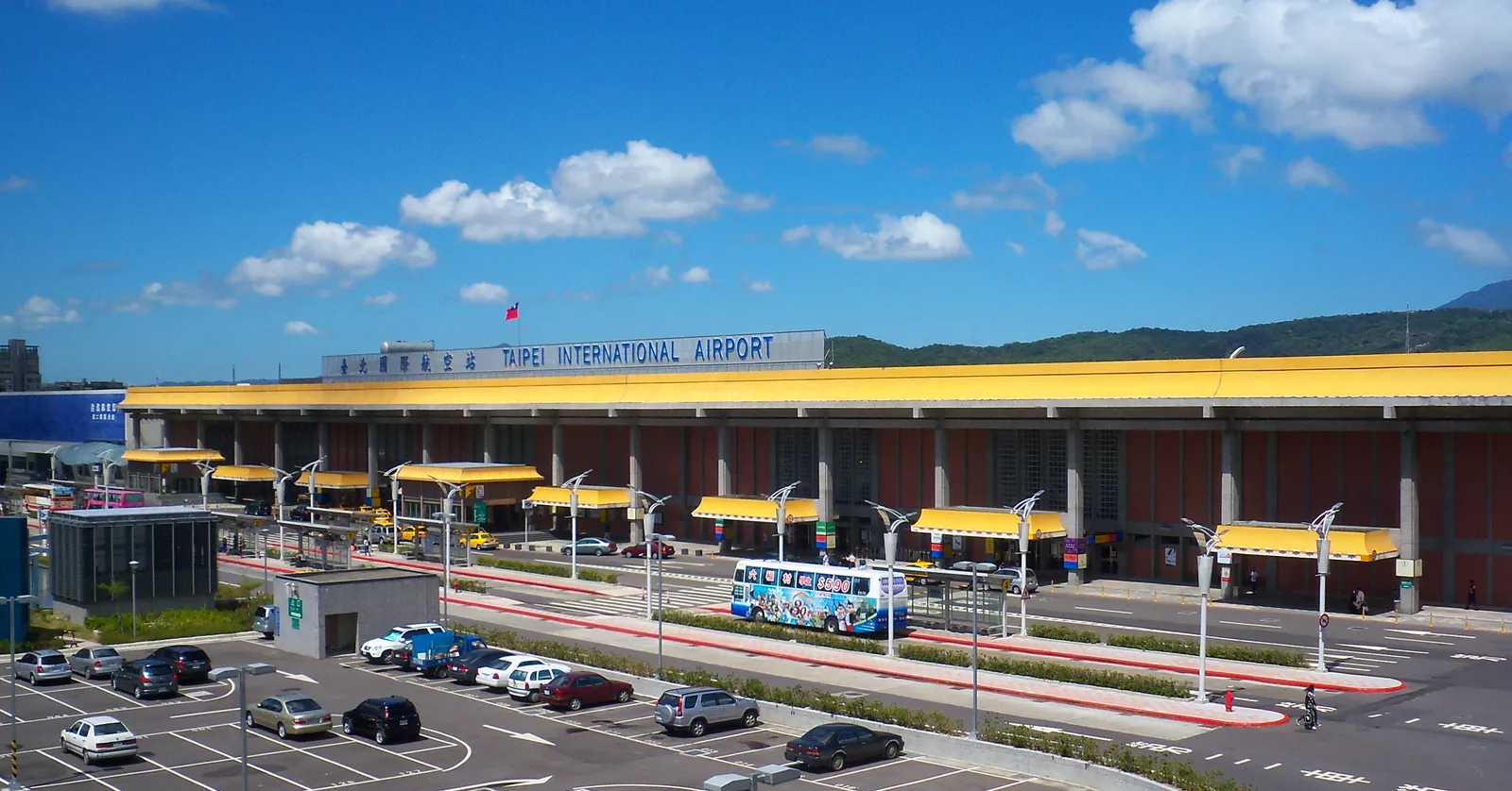 The long low terminal building of Taipei Songshan Airport under a row of yellow canopies, with cars in the forecourt and green hills behind the roofline
