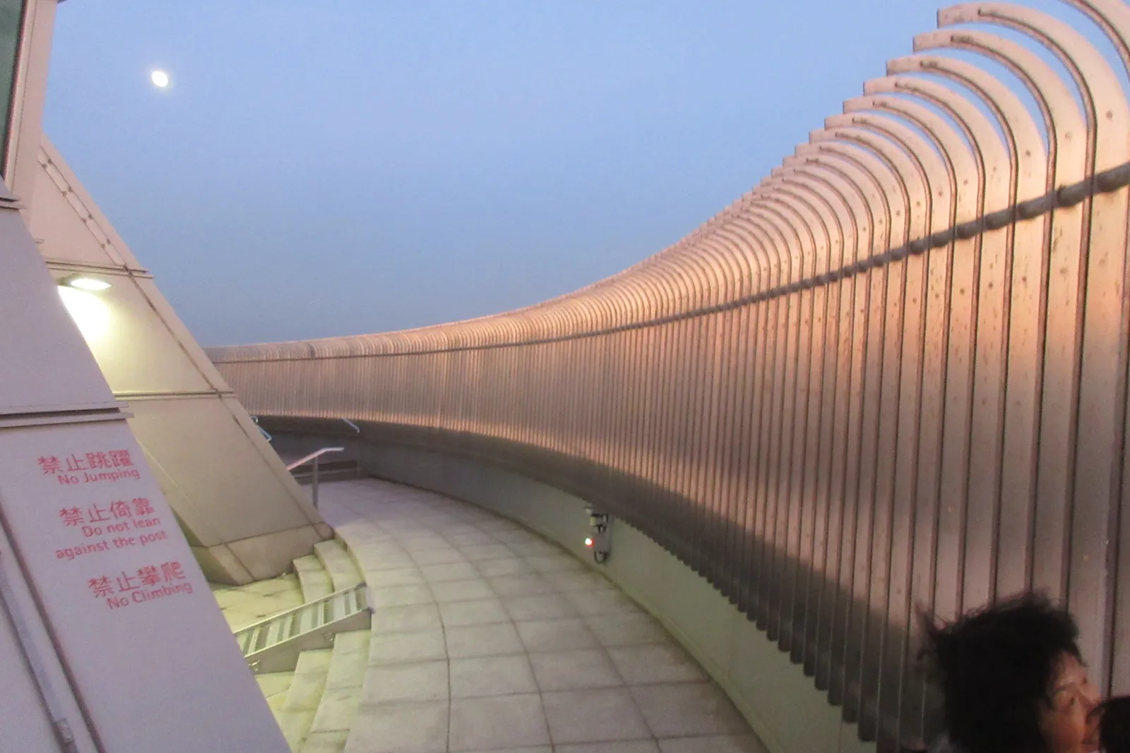 The open-air terrace on the 91st floor of Taipei 101, a curving wall of copper-coloured metal louvres shielding the walkway, with the moon in the sky above it