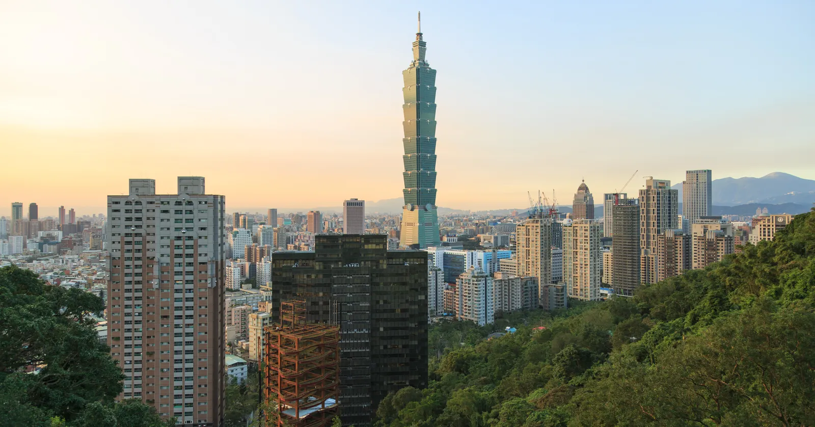 Taipei 101 rising above the apartment blocks and office towers of Xinyi at sunset, seen from a wooded slope with hills on the horizon