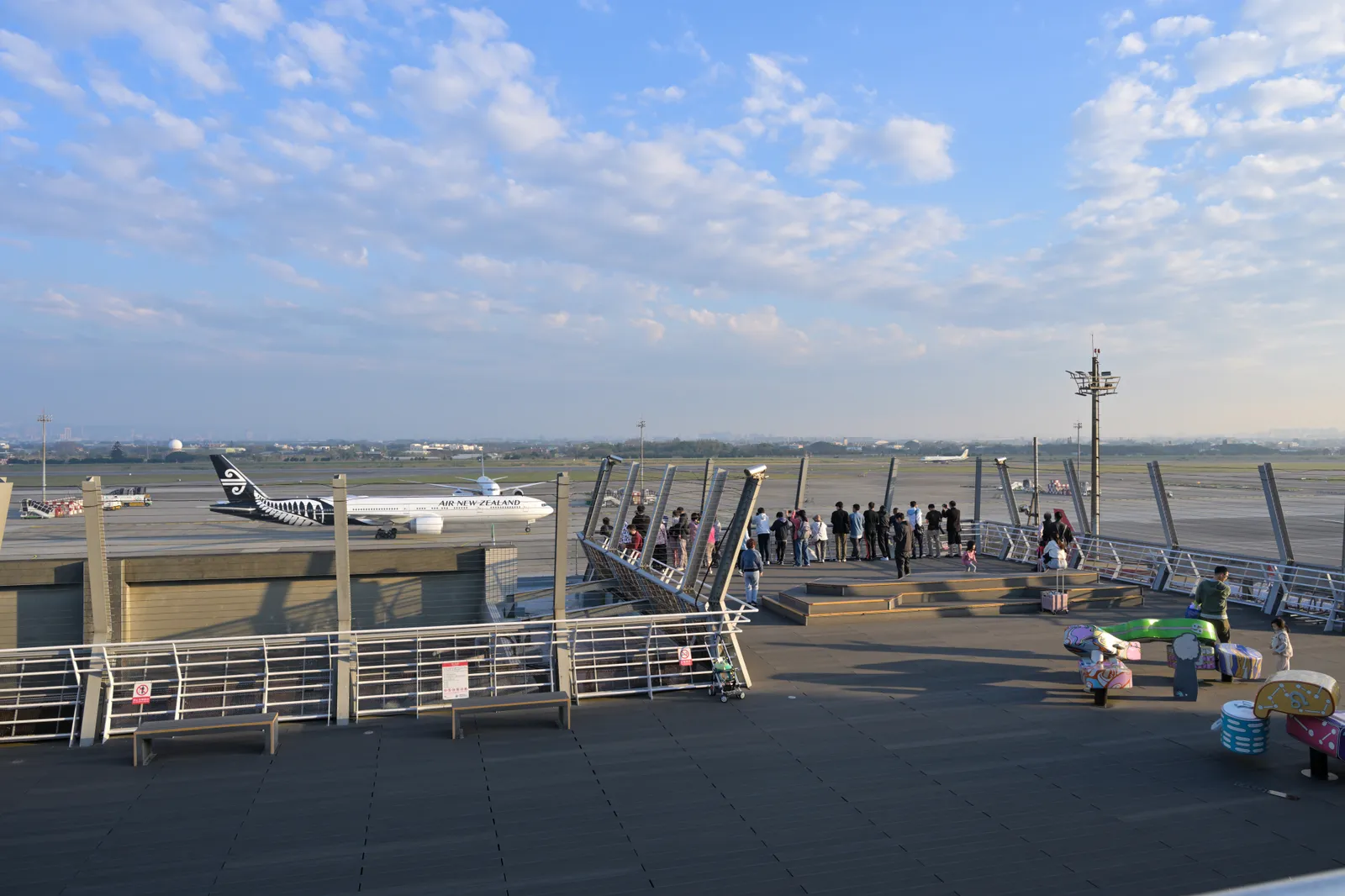 People standing along the railing of the open-air observation deck at Taoyuan Airport, looking out over the apron with an airliner parked at a stand behind them