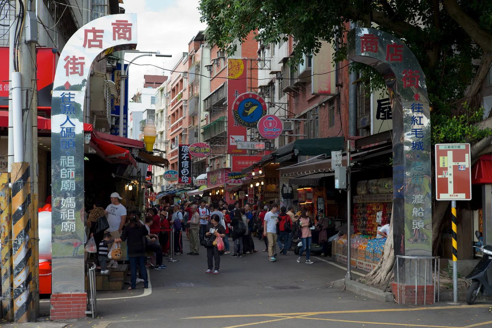 The entrance arch over Tamsui Old Street in New Taipei, with shopfronts, awnings and vertical signs crowding both sides of the lane and shoppers walking down the middle