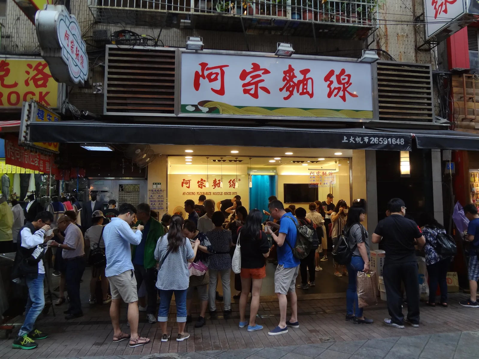 The red and white shopfront of the Ay-Chung noodle stall on Emei Street in Taipei, with a crowd of customers standing in the street outside eating from bowls