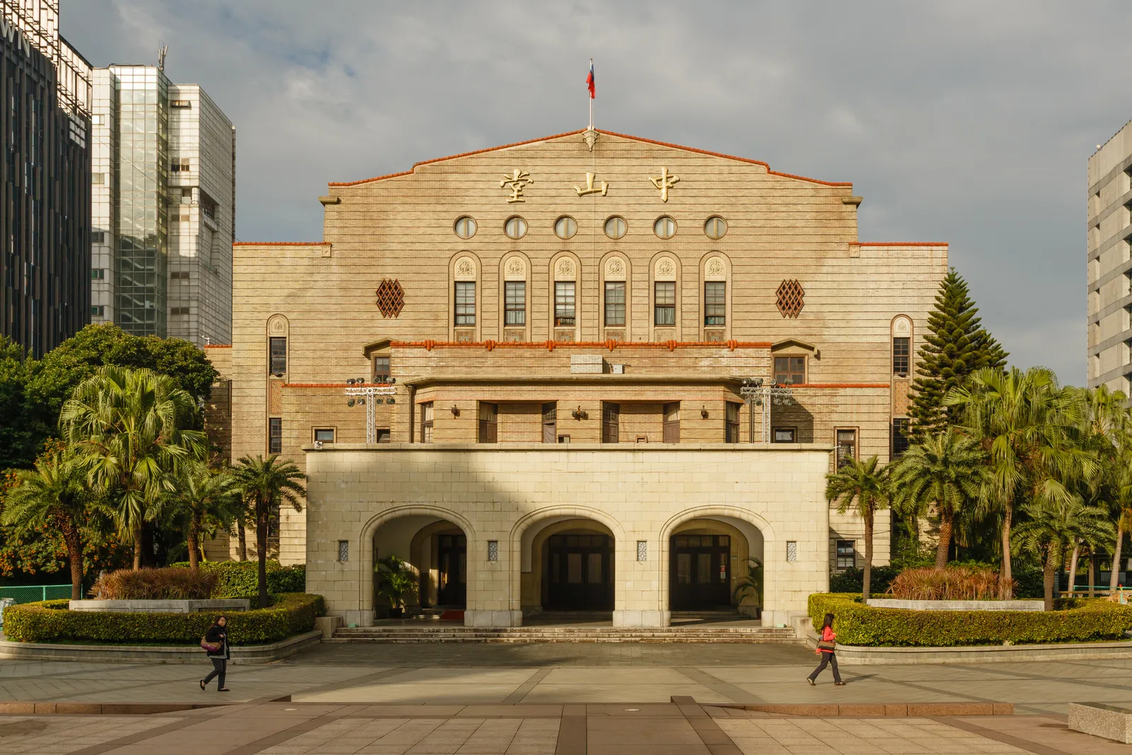 The pale brick facade of Zhongshan Hall in Taipei, a 1930s civic building with three tall arched entrances at ground level and a row of windows above them