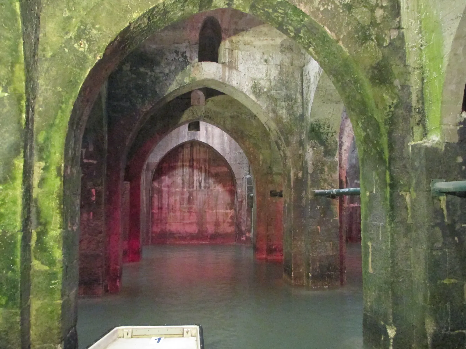 Inside the Pool of Arches in Ramla, pointed stone arches springing from square piers into still green water, moss on the lower stonework and the white prow of a rowing boat in the foreground