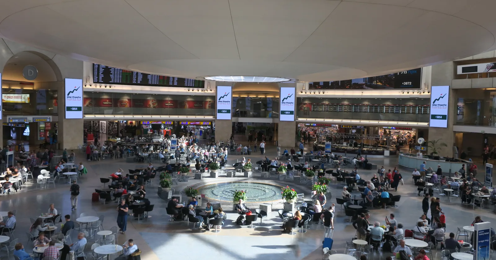 The star-shaped duty-free rotunda at Ben Gurion Airport seen from the balcony above, with a circular stone fountain in the middle of the floor and travellers sitting at cafe tables all around it
