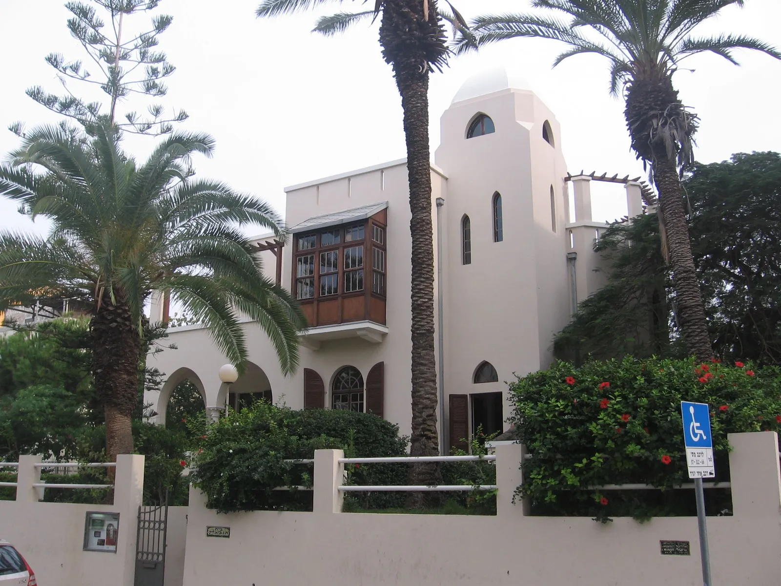 Bialik House in Tel Aviv, a white 1920s villa with a domed corner tower, arched windows and a projecting wooden bay, half hidden behind date palms and a low garden wall