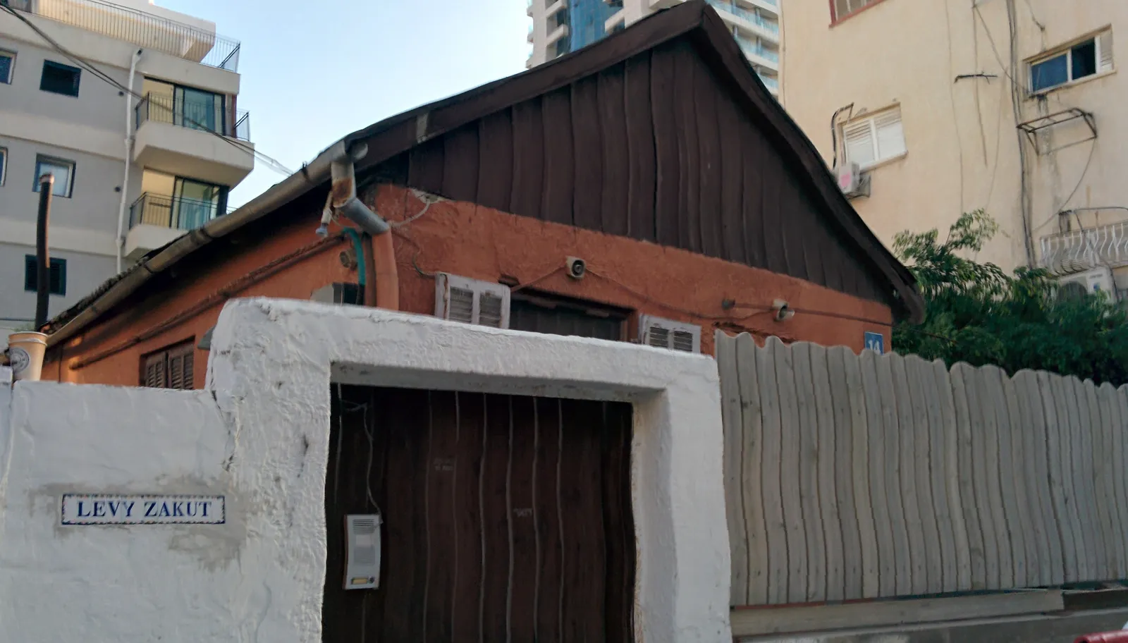 A single-storey house with a pitched tin roof and a brown wooden door behind a whitewashed wall in Kerem HaTeimanim, Tel Aviv, with new apartment towers rising directly behind it