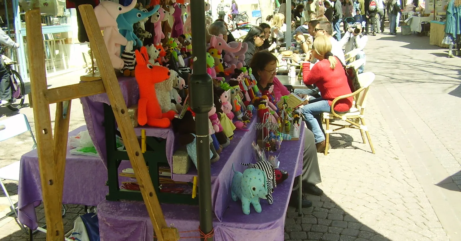 Stalls at the Nahalat Binyamin craft fair in Tel Aviv, hand-sewn cloth animals hanging from a frame above purple-draped tables, with green awnings and more stalls running down the pedestrian street behind