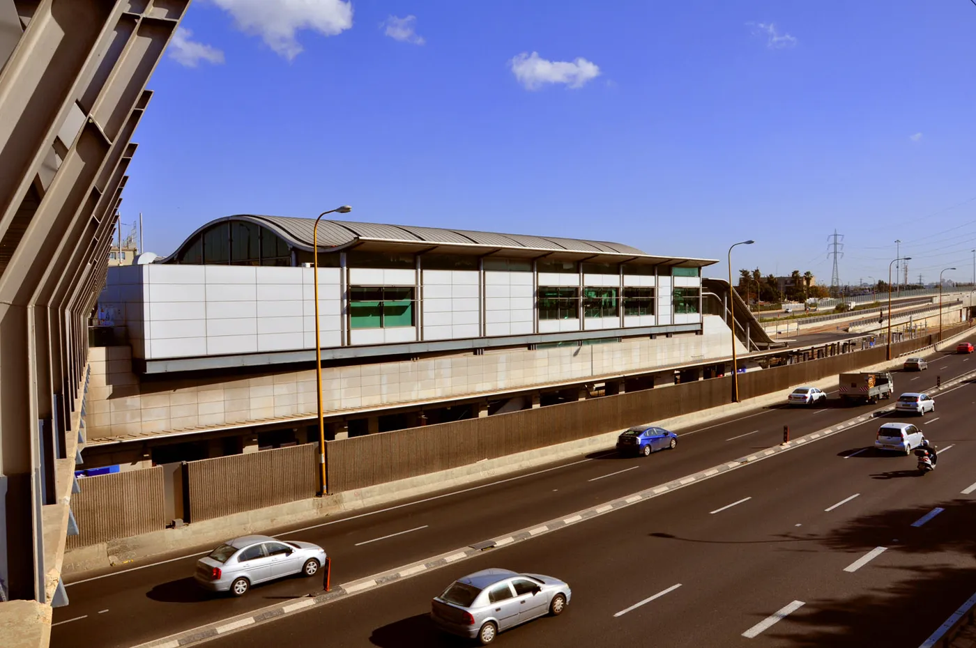 Tel Aviv HaHagana station seen from above, a long glass and steel platform building sitting in the middle of the Ayalon motorway with traffic running past on both sides