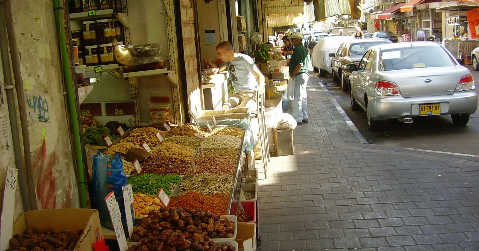 A spice shop on Levinsky Street in Tel Aviv with open trays of dried fruit, nuts and pulses spilling out onto the pavement under a shaded arcade, cars parked along the kerb beside them