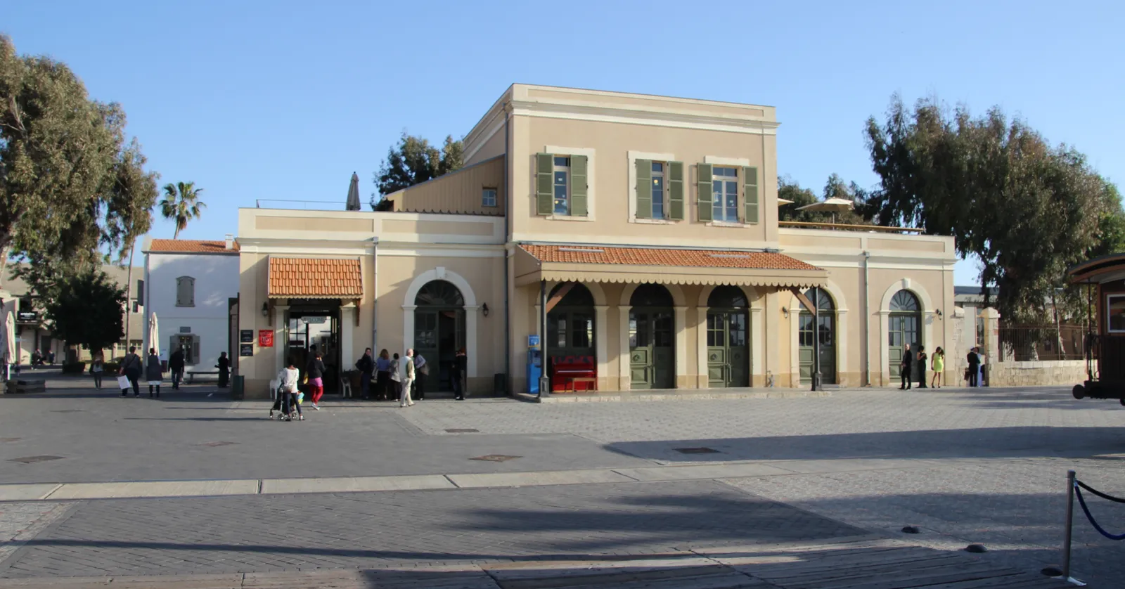 The restored Ottoman-era Jaffa railway station building in Tel Aviv, a pale two-storey block with green shutters and a red-tiled arcade of awnings along its front, standing across an open paved plaza