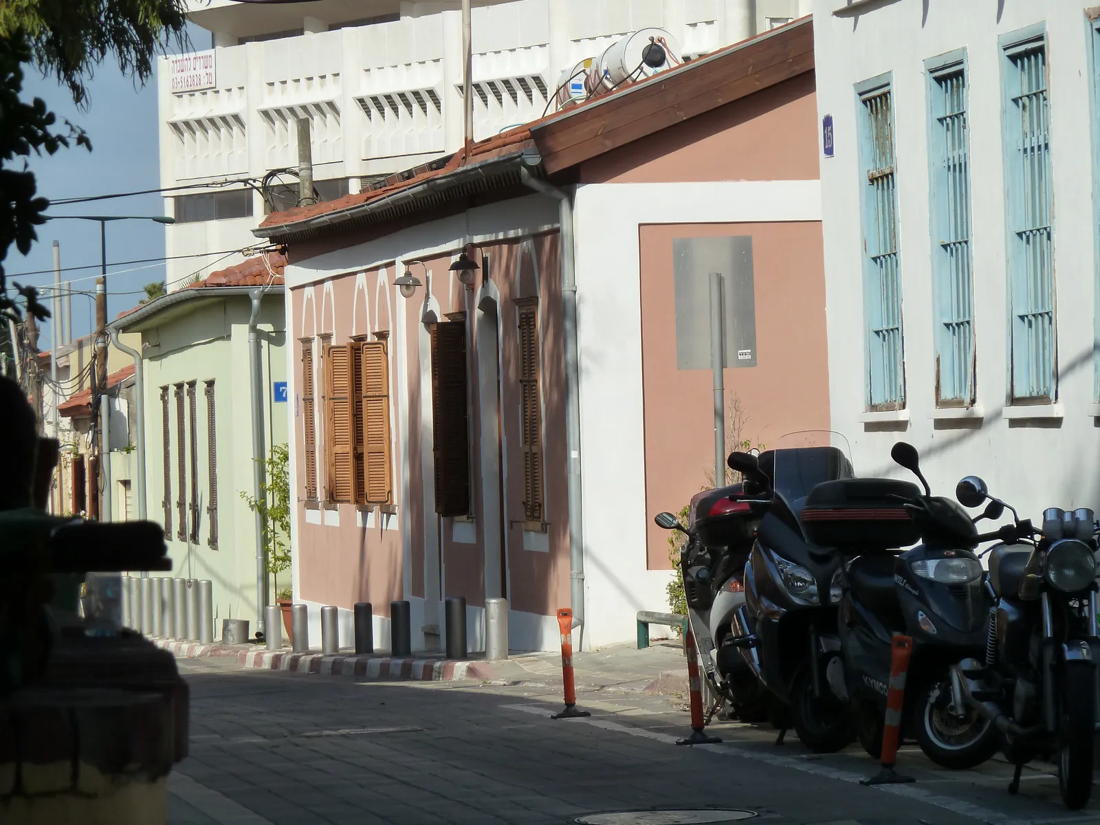 A lane in Neve Tzedek, Tel Aviv, with low one-storey houses in pink, green and white, wooden shutters and tiled eaves, and scooters parked nose-in along the kerb