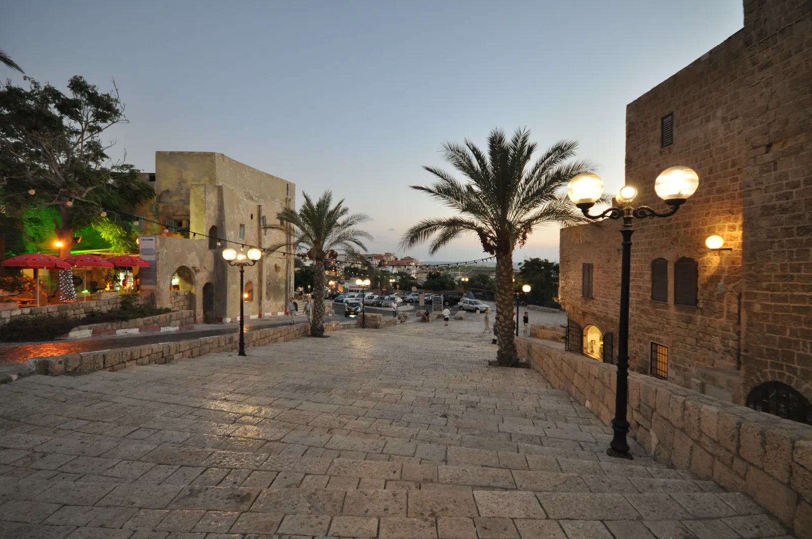 Kedumim Square in Old Jaffa at dusk, broad pale flagstones running between sandstone walls, ornate black lamp posts lit along one side and palm trees standing over the far end