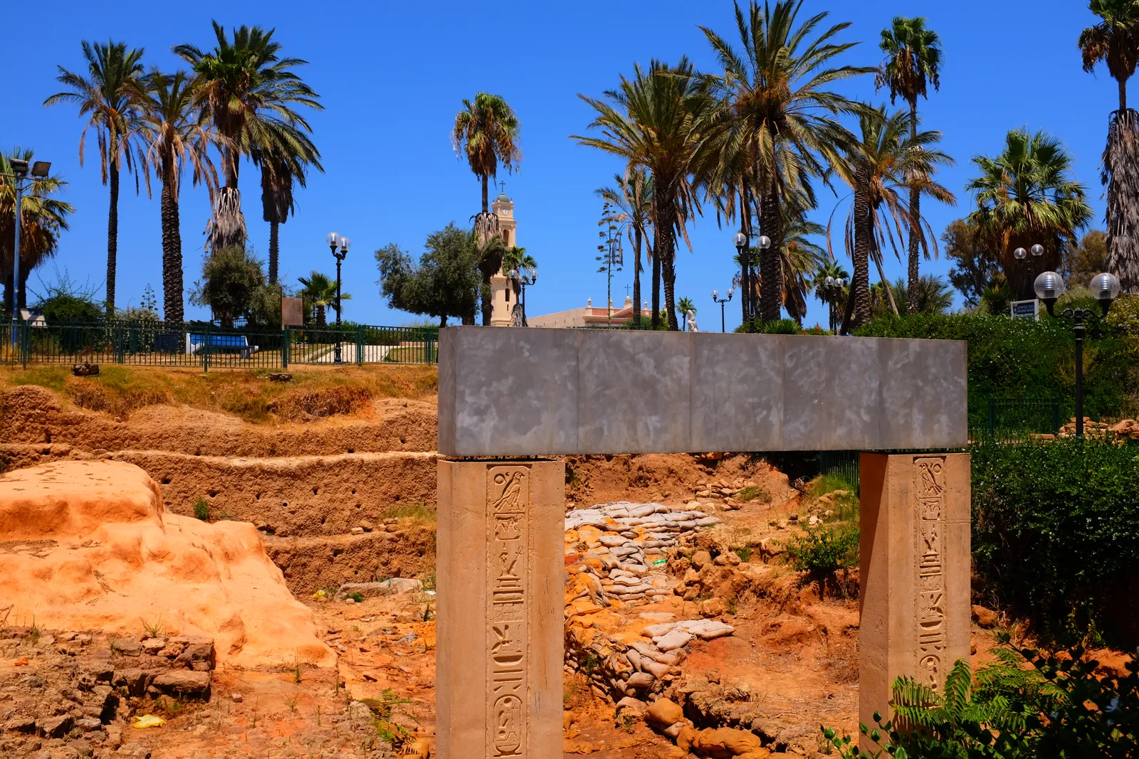 The reconstructed Egyptian gateway in HaPisga Garden, Old Jaffa, two inscribed stone jambs carrying a plain lintel above an excavated trench of red earth and stone, with palms and a bell tower behind