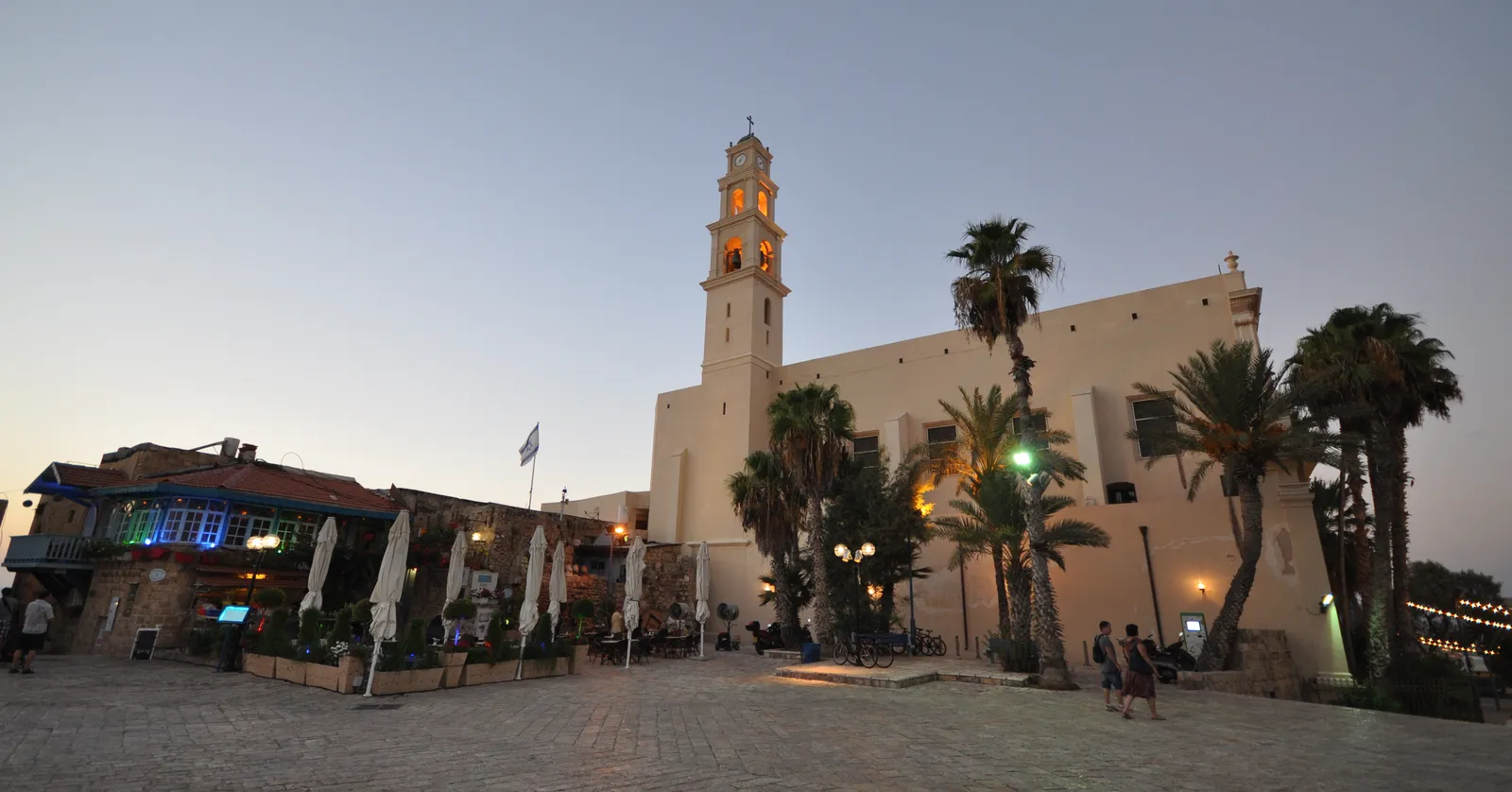 St Peter's Church in Old Jaffa at dusk, a tall pale bell tower lit gold above a long buttressed nave, with palms and lit cafe terraces along the square in front of it