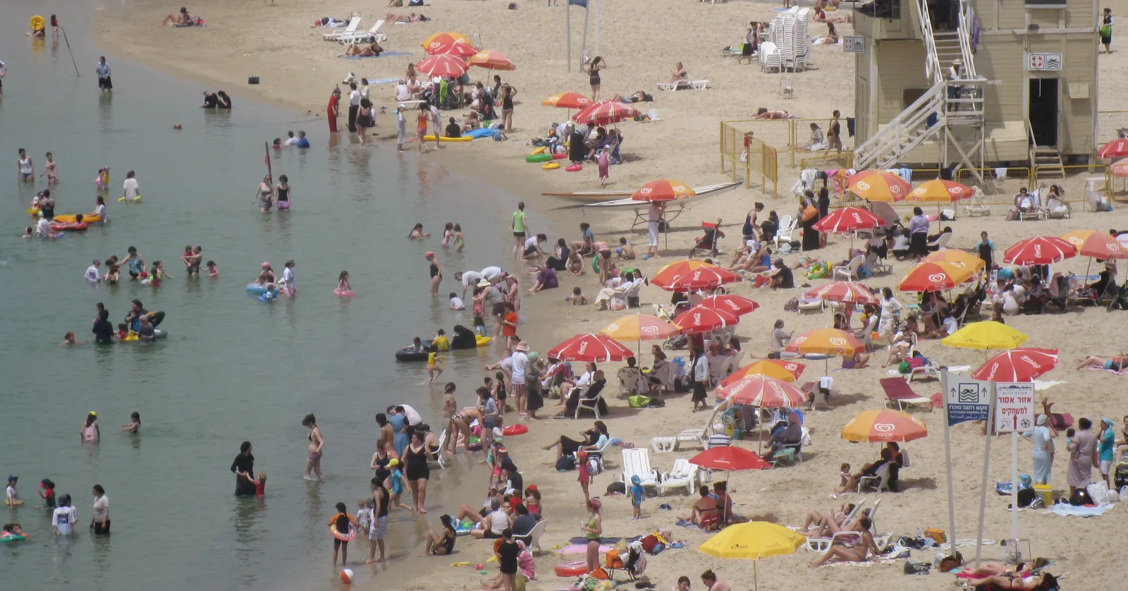 Metzitzim Beach in Tel Aviv packed with red and yellow parasols, bathers standing in shallow flat water on the left and a lifeguard tower on the sand at the right